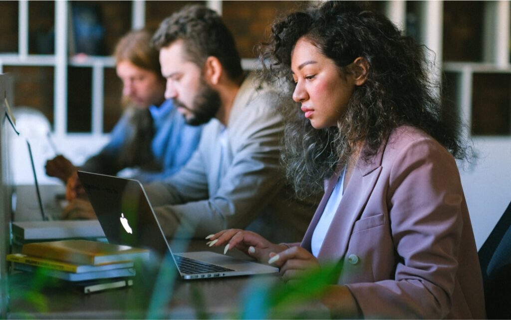 une femme en veste rose devant son ordinateur portable sur un bureau, deux hommes collaborateurs en arrière plan, girarin industriel