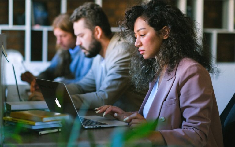 une femme en veste rose devant son ordinateur portable sur un bureau, deux hommes collaborateurs en arrière plan, girarin industriel